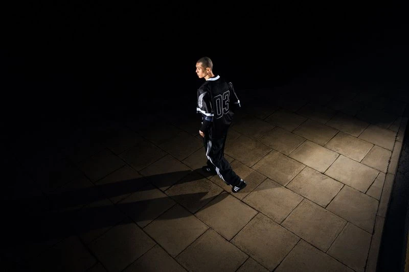 An adult male model with short hair wearing a dark sports outfit stands on a brick sidewalk with shadows in a nighttime urban setting.