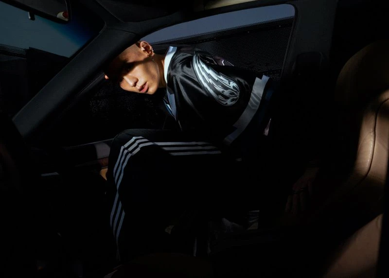 Man in a car interior wearing an Adidas track suit and jacket, creating a dark, moody, and enclosed atmosphere with limited light.