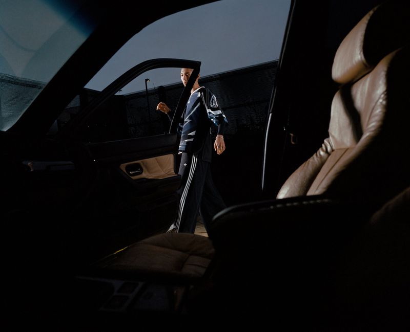 Adult posing near a black car, wearing a navy and white tracksuit with triple stripes, with a leather interior car seat visible.