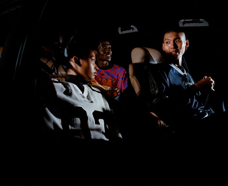Three people inside of a vehicle, the focal point on a man with an open mouth and earrings, alongside two other passengers in dim lighting.