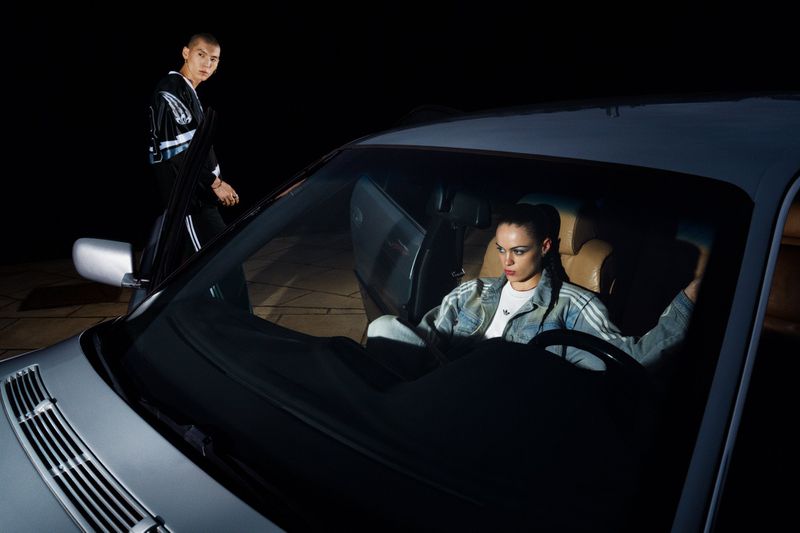 Young woman sits in car at night with man standing nearby in Adidas tracksuits. Headlights illuminate the scene in an urban setting.