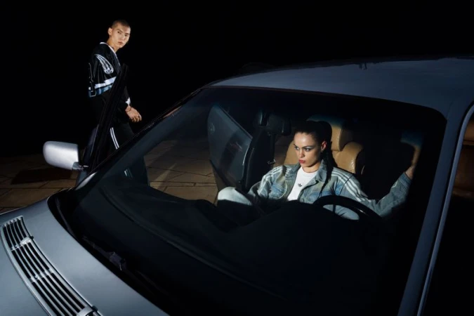 Young woman sits in car at night with man standing nearby in Adidas tracksuits. Headlights illuminate the scene in an urban setting.