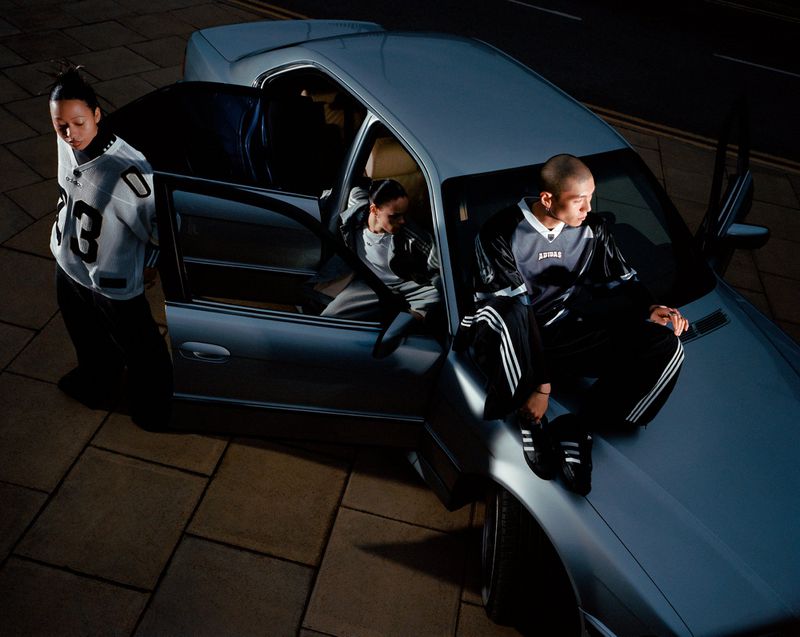 Three people and a silver sedan car are photographed at night. One person sits on the hood, one in the car, and one stands by the open door.