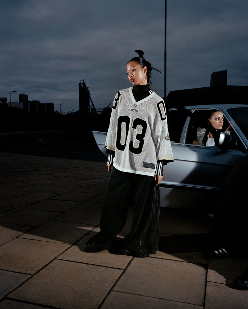 A young Asian woman wearing a football jersey stands next to a car with another person inside, with buildings in the background at dusk.