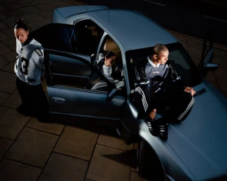 Three models wearing sportswear posing around a silver sedan parked on tiled pavement with open doors.
