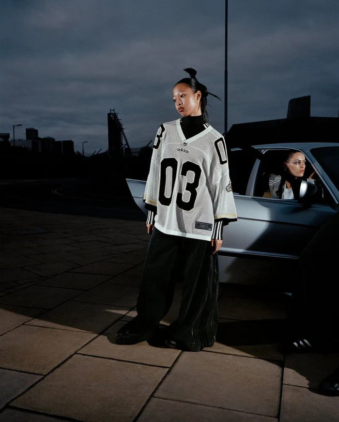 Model standing outdoors wearing an oversized white jersey next to a silver car at dusk for Adidas