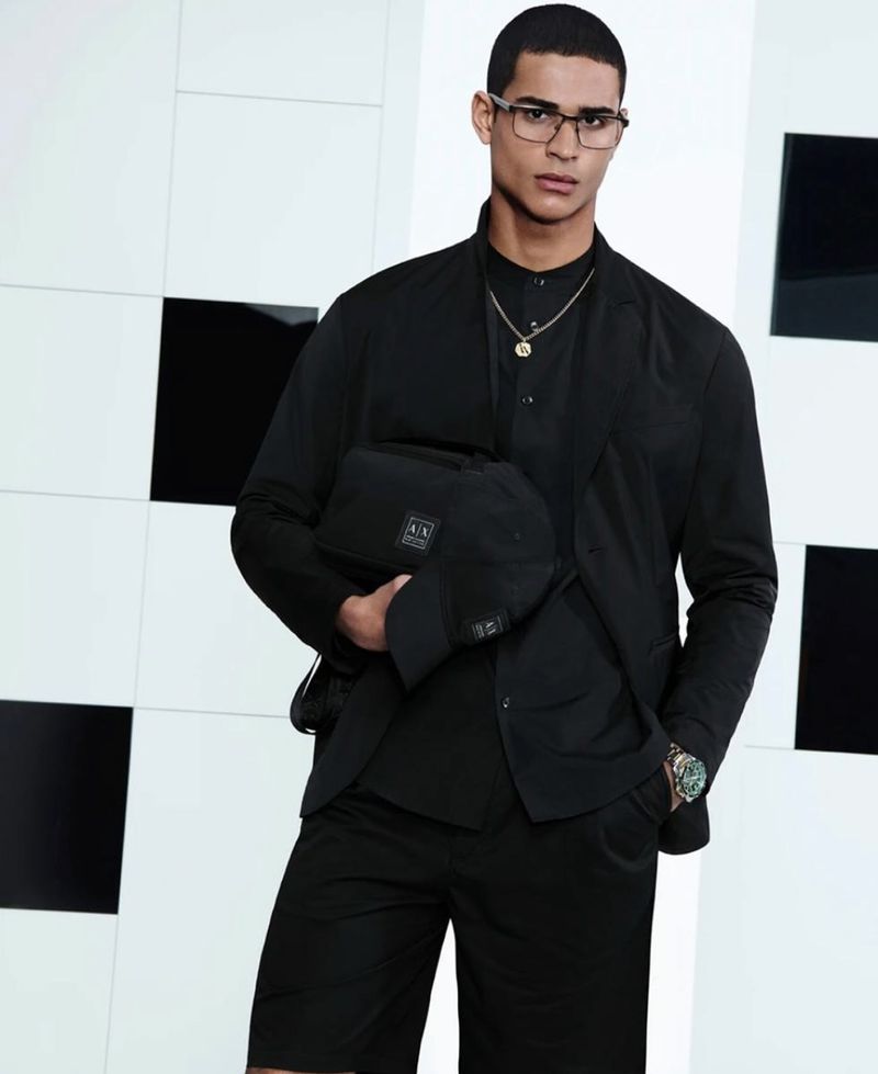Man wearing a black suit and holding a black cap and bag against a patterned background.