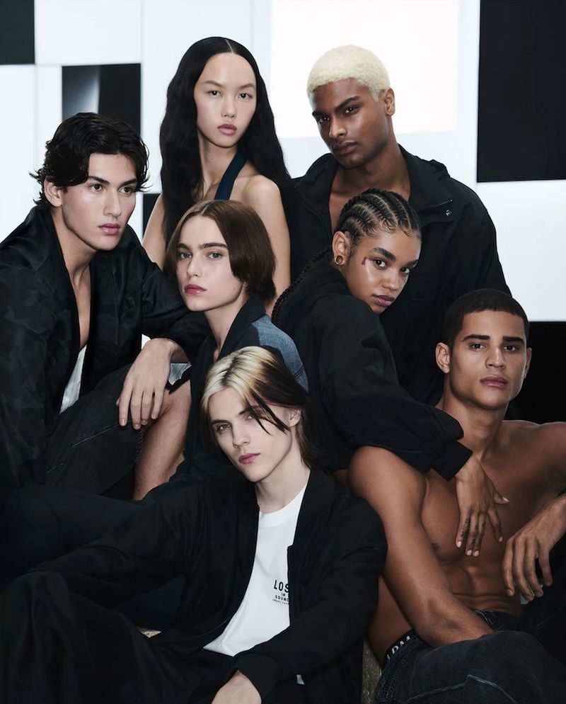 Seven diverse individuals are indoors in a group shot wearing black jackets, jeans, and a white t-shirt against a black and white geometric backdrop.