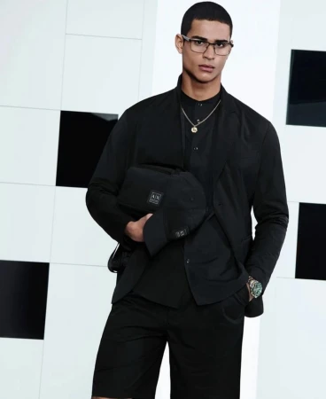 Man wearing a black suit and holding a black cap and bag against a patterned background.