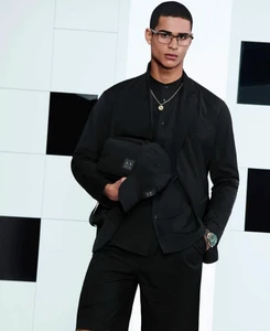 Man wearing a black suit and holding a black cap and bag against a patterned background.