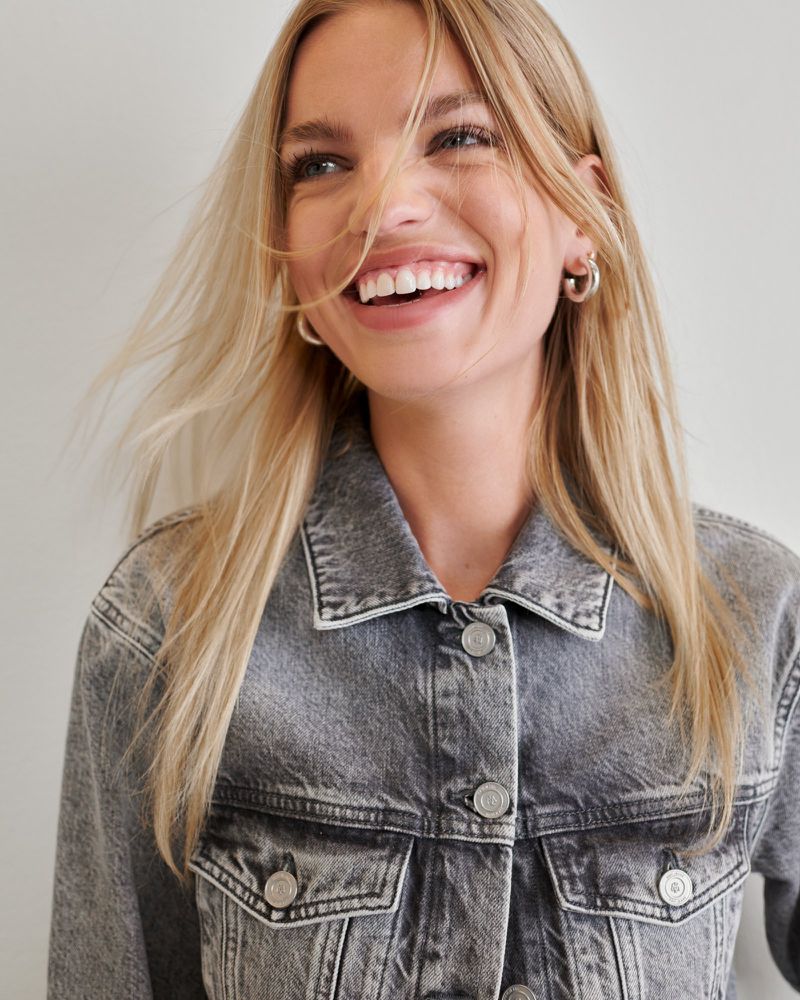Smiling blond female model wearing a grey denim jacket against a neutral backdrop