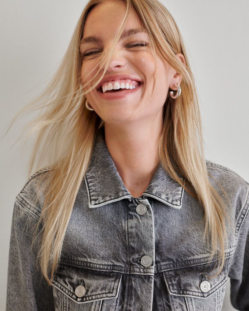 Smiling blonde woman in a grey denim jacket against a white backdrop, capturing a cheerful and casual fashion moment.