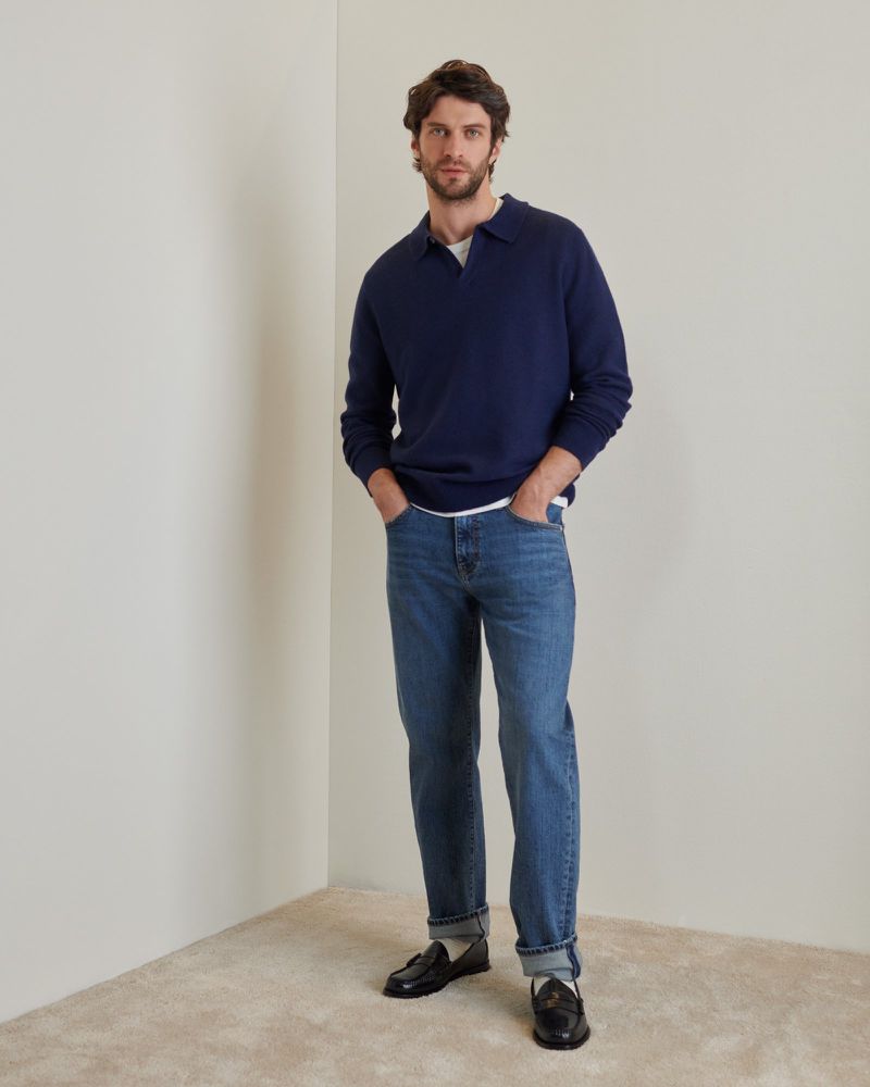 A stylish adult male in a navy sweater and jeans stands indoors. He's captured in a studio shot with a neutral background.