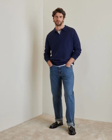A stylish adult male in a navy sweater and jeans stands indoors. He's captured in a studio shot with a neutral background.