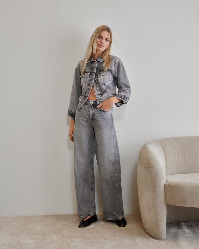 Blonde woman in a grey denim jacket and wide-leg jeans poses indoors against a wall next to a modern, cream-colored chair.