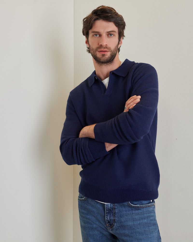 A man in a blue collared sweater and jeans stands indoors with arms crossed, exuding casual confidence in a simple, well-lit setting.