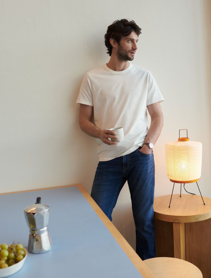 Man in white t-shirt and jeans leans against wall, holding mug. Side table with lit lamp and bowl of grapes.