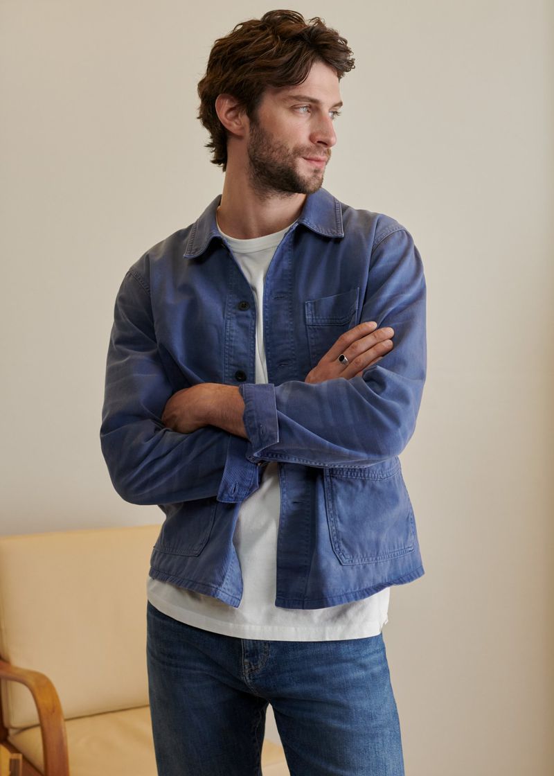 A medium shot of a man in a blue jacket and jeans, looking to the side in a neutral indoor setting with natural lighting. Fashion.