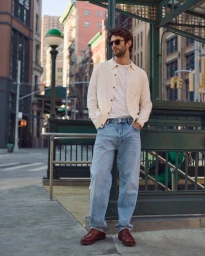 Man wearing a cream button-down shirt and light wash jeans standing beside a subway entrance on a city street.