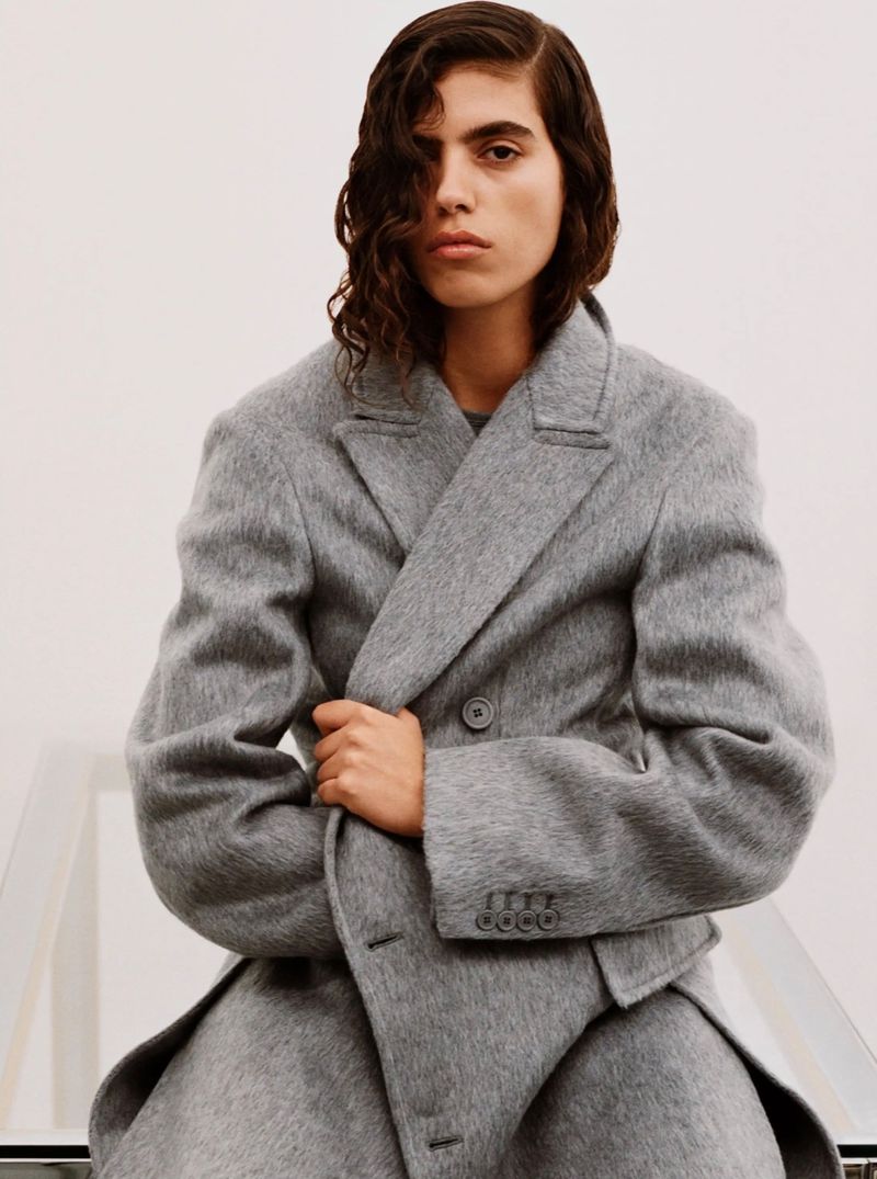 Close-up of a person with long wavy hair wearing a grey wool overcoat. The individual is sitting in a light coloured interior.
