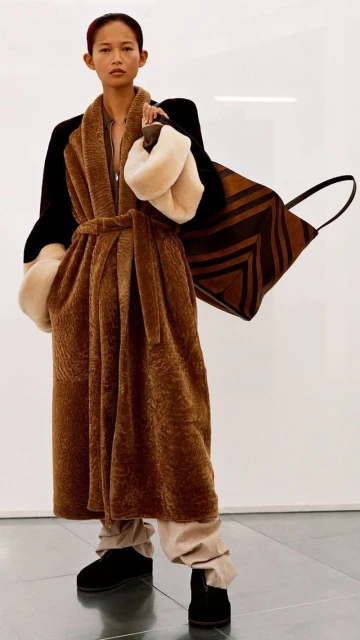 Fashion model poses in a brown faux-fur robe coat with a geometric print tote bag, indoor setting, promoting fall-winter collection.