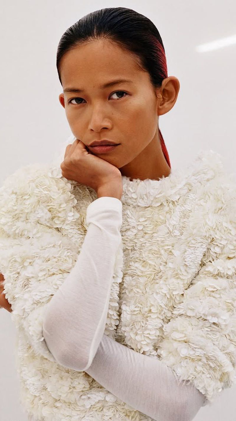 Portrait of an Asian woman with dark hair styled back, posing indoors wearing a textured white top and long white sleeves.