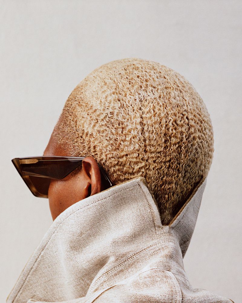 Close-up studio shot of a Black adult female model with short blonde curly hair wearing sunglasses and a light brown jacket.