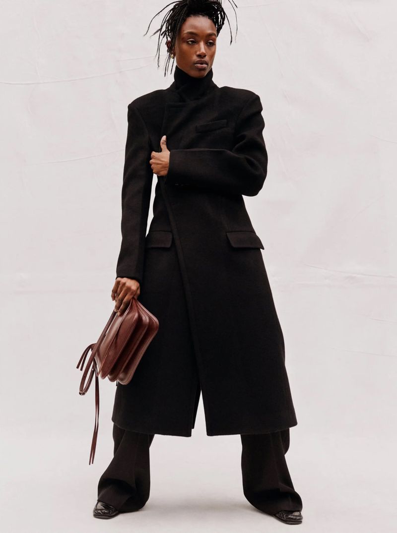 A Black woman in a long black coat and wide-legged pants holds a brown leather purse against a white backdrop in a fashion studio shoot.