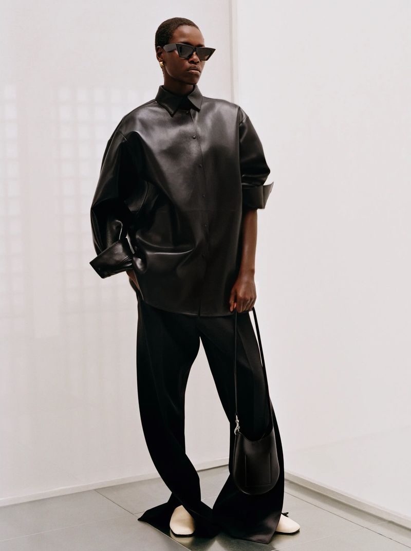 A woman in a black leather shirt, black pants, and white shoes holds a small black bag in a minimalist studio setting.