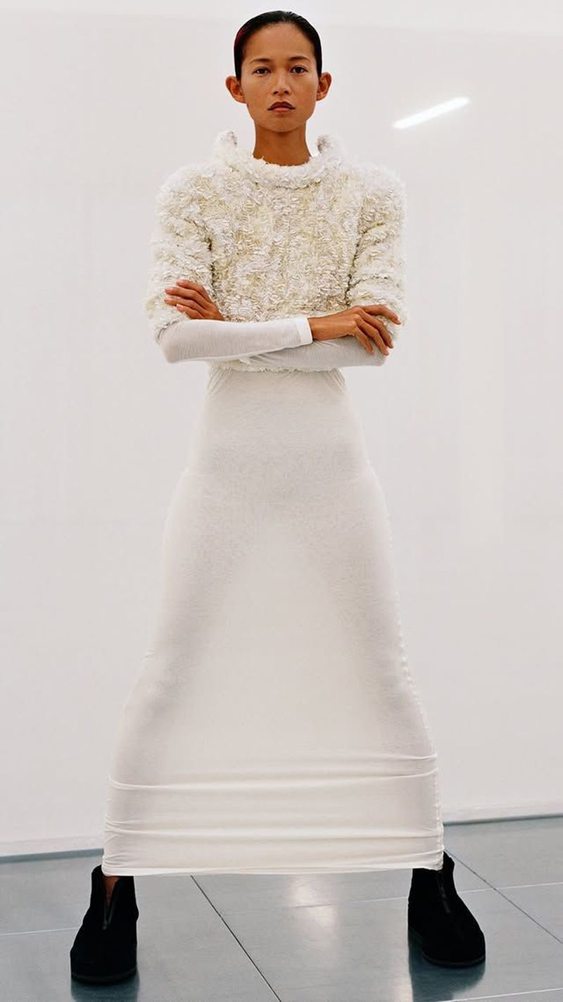 Full shot of a slender, Asian-appearing woman in a textured white top, long white skirt, and black shoes, posing against a white background.