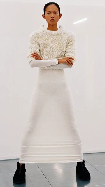 Full shot of a slender, Asian-appearing woman in a textured white top, long white skirt, and black shoes, posing against a white background.