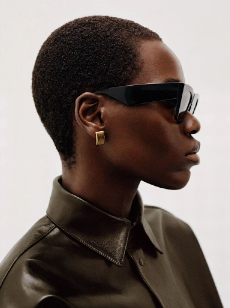 Profile portrait of a model wearing black sunglasses, a gold earring, and a dark olive leather shirt against a white backdrop.
