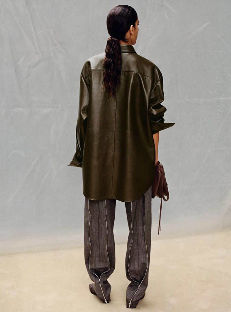 Back view of a woman with a ponytail wearing an olive green faux leather shirt and grey trousers with a dark red handbag.