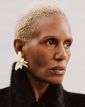 Close-up portrait of an adult Black woman with short bleached blonde hair wearing a dark coat and cream-colored statement earrings.