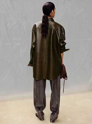 Model seen from behind wearing an olive green leather shirt and grey trousers against a grey backdrop