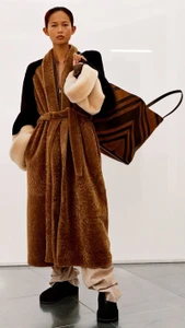 Model wearing a brown belted coat and carrying a large striped tote bag against a white studio background.