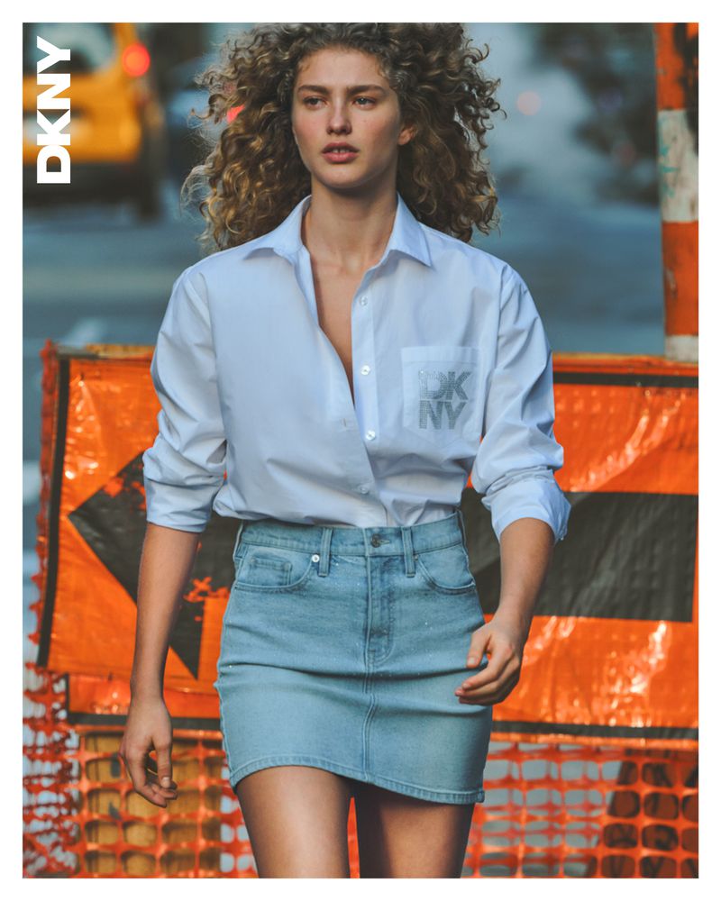 Fashion shot of a woman in a white button-down shirt and jean skirt on a city street, posing near construction barriers.