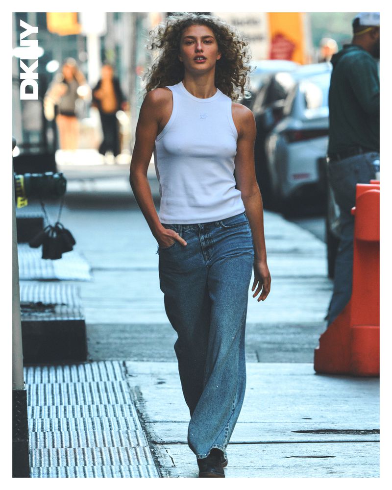 Model with curly hair wearing a white tank top and blue jeans walking down a city street for the DKNY Spring 2025 Campaign
