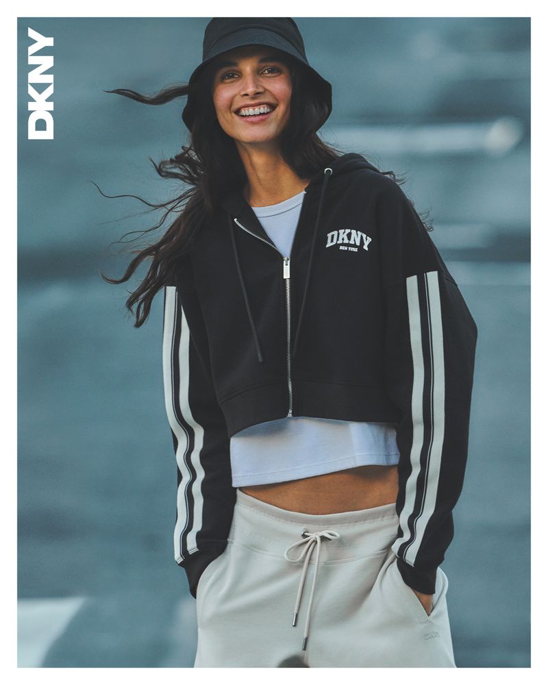 Smiling woman in bucket hat and DKNY athleisure wear poses against a blue background, showcasing casual fashion and brand appeal.