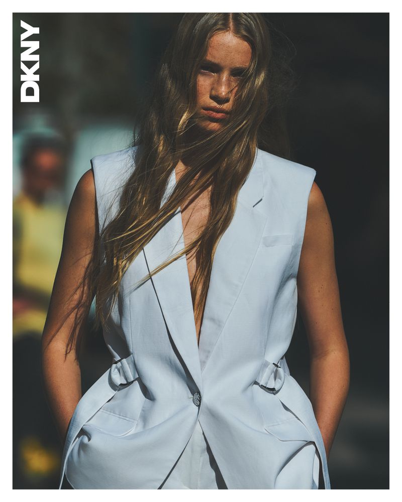 Fashion editorial shot of a blonde woman wearing a light blue suit vest and facing forward, with a second person blurred in the background.