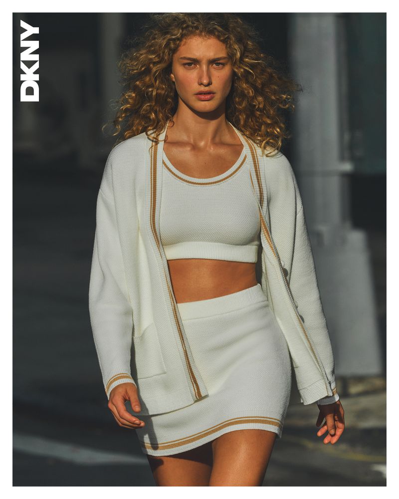Fashion shot of a young adult woman with curly hair wearing a white two-piece knit set and a white cardigan in an urban setting.