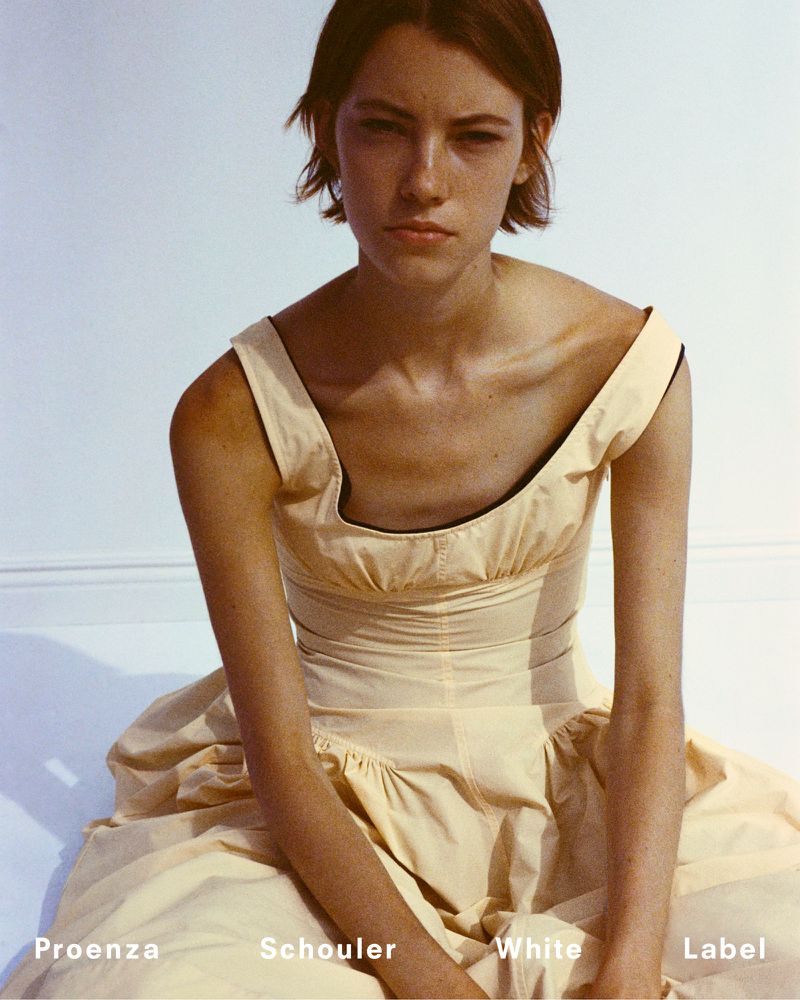 A fair-skinned woman with short brown hair poses in a light-yellow dress against a white backdrop, with Proenza Schouler branding visible.