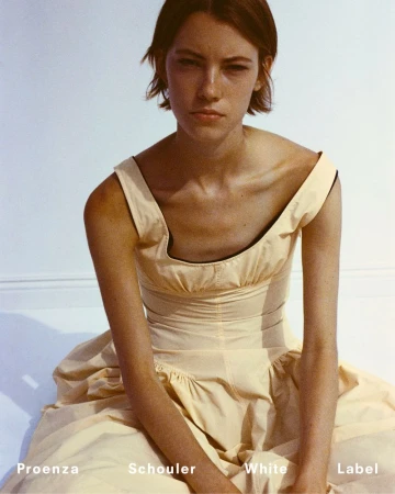 A fair-skinned woman with short brown hair poses in a light-yellow dress against a white backdrop, with Proenza Schouler branding visible.