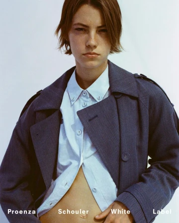 A woman with short hair poses in a light blue shirt and dark blue jacket against a white background in a fashion-style photo.
