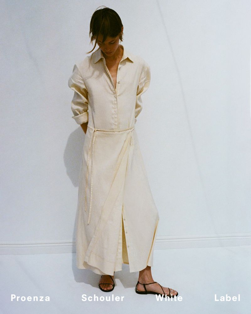 A woman poses in a long, light-colored dress with sandals against a white backdrop, showcasing a fashion brand label.