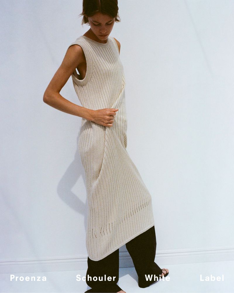 Indoor shot of a slender woman in a cream-colored, knit dress, black pants, and bare feet against a plain white background.