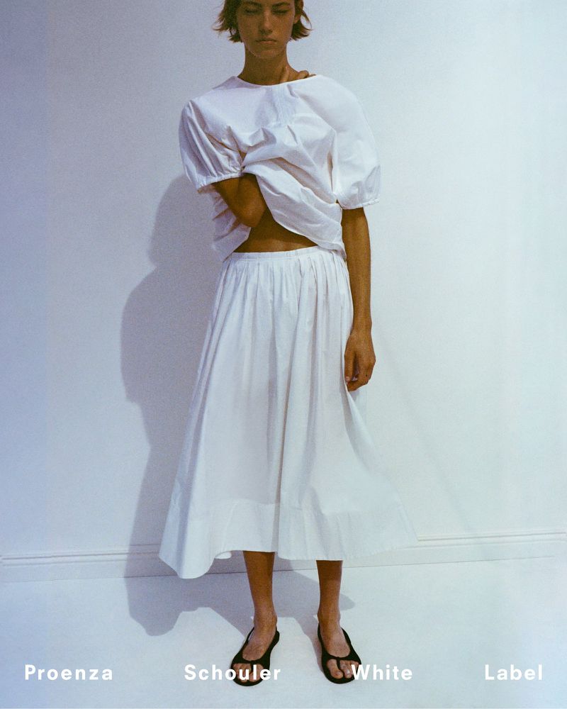 A woman in a white, puffy-sleeved blouse and flowing skirt poses in black sandals against a white wall in a fashion-forward studio shot.