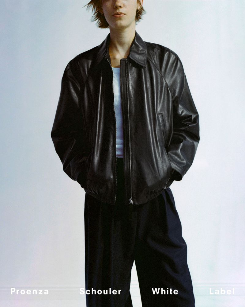 Model wearing a black leather jacket over a white t-shirt and dark trousers against a plain white studio backdrop.
