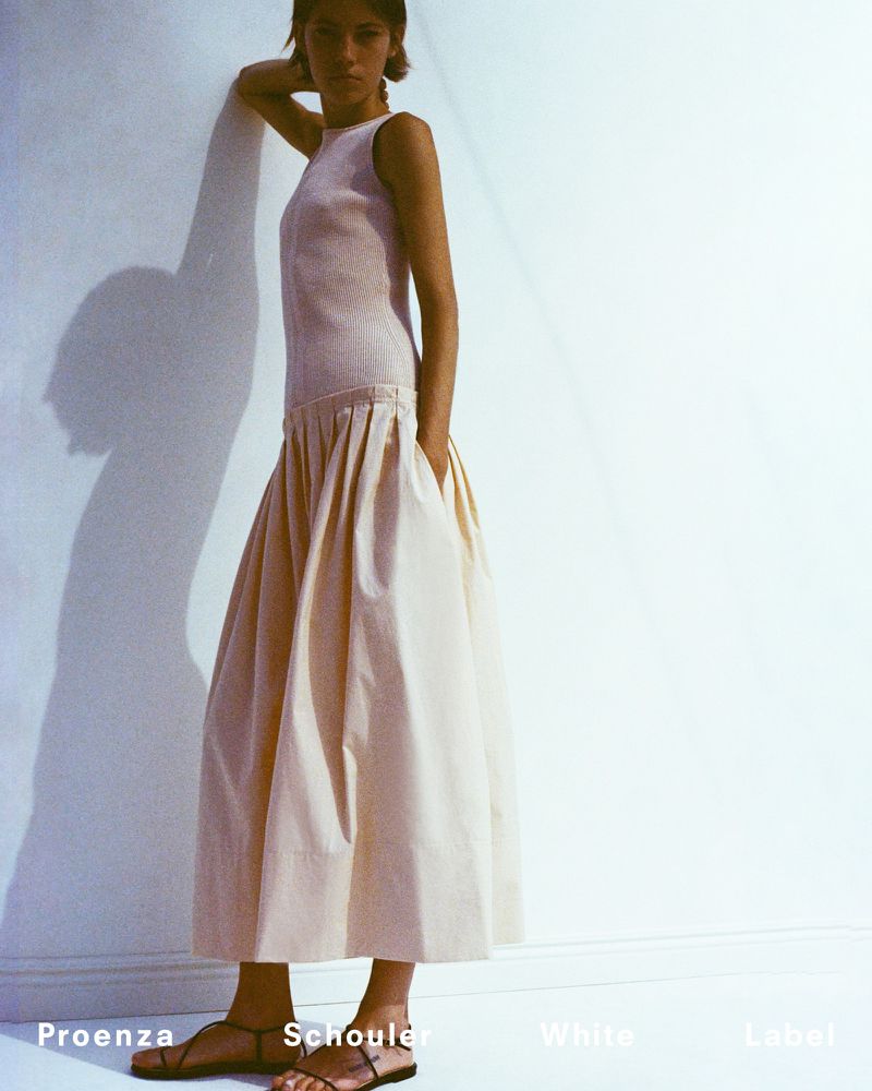 A woman models a cream-colored dress, featuring a drop waist and full skirt, against a light background with shadow detail. She wears flat sandals.