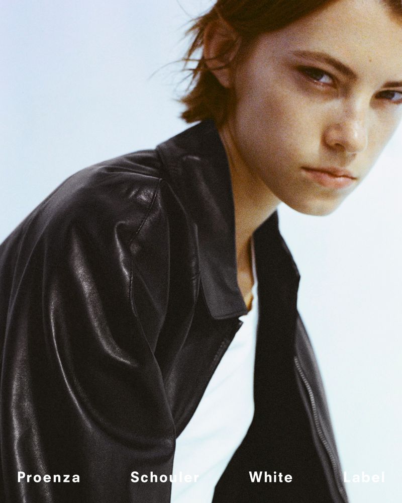 Close-up studio portrait of an adult woman posing with a black leather jacket, wearing a white top, against a pale blue background.